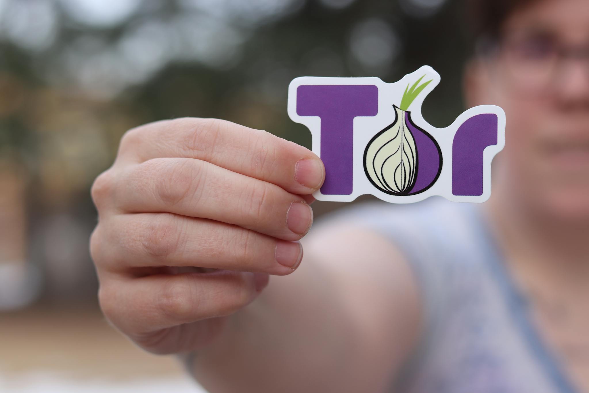A close-up of a person holding a Tor browser logo sticker with a blurred outdoor background.